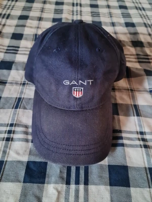 Mörkblå keps från GANT - Snygg mörkblå keps från GANT med broderad logga och sköld framtill. Klassisk böjd skärm och justerbar rem baktill. Tillverkad i bomullstyg som ger en avslappnad och sportig look.