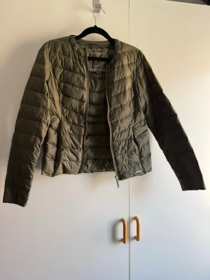 Olivgrön pufferjacka från Ilse Jacobsen - Snygg olivgrön pufferjacka från J.Lindeberg med quiltad design och rund halsringning. Jackan har dragkedja framtill och är tillverkad i lätt, vadderat material som håller värmen. Perfekt för kalla dagar och enkel att styla till olika outfits. Endast provad katt finns i hem!