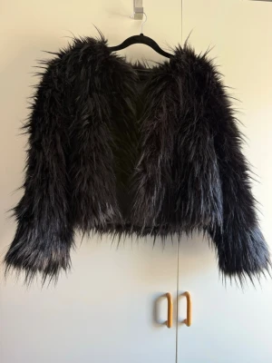 Svart fluffig fuskpälsjacka - Säljer en svart, kort fuskpälsjacka med lång, fluffig päls och öppen front. Jackan har en boxig passform och långa ärmar. Perfekt för dig som vill sticka ut med en trendig och mysig look under kalla dagar. Endast testad, katt finns i hem!
