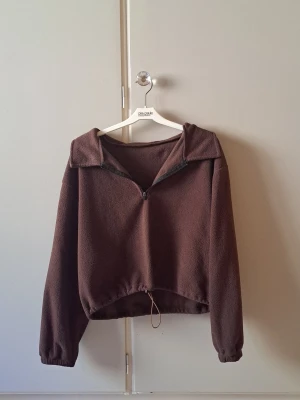 Brun fleece-tröja med dragkedja - Mysig brun fleece-tröja med halv dragkedja framtill och krage. Tröjan har långa ärmar, resår vid ärmslut och justerbar dragsko i nederkant. Perfekt för lager-på-lager och chill dagar. O typ friluftsliv eller äventyrslook. Storlek XS/S
