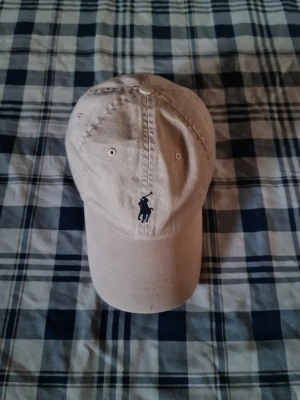 Beige keps från Polo Ralph Lauren - Snygg beige keps från Polo Ralph Lauren i 100% bomull. Klassisk modell med böjd skärm och broderad marinblå logga framtill. Perfekt accessoar för en avslappnad och stilren look.