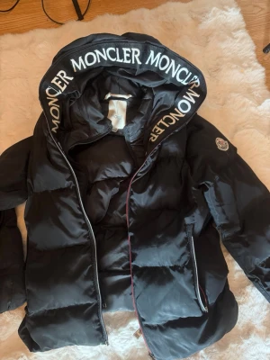 Svart dunjacka från Moncler - Snygg svart dunjacka från Moncler med stor huva och tydlig logga på ärmen. Jackan har dragkedja, två fickor och Moncler-tryck på huvkanten. Perfekt för kalla vinterdagar och riktigt varm tack vare dunfyllningen. Storlek 11/12 år. Dragkedjan är lite trasig men enkelt att fixa.