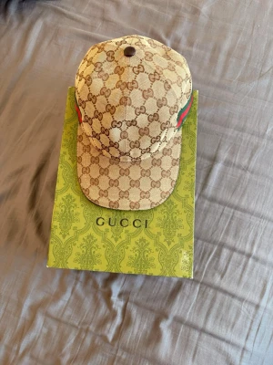 Beige keps från Gucci med GG-mönster - Snygg beige keps från Gucci med klassiskt GG-monogram i brunt över hela kepsen. Kepsen har gröna och röda detaljer på sidorna och justerbart spänne bak i brunt läder. Perfekt accessoar för en trendig look.