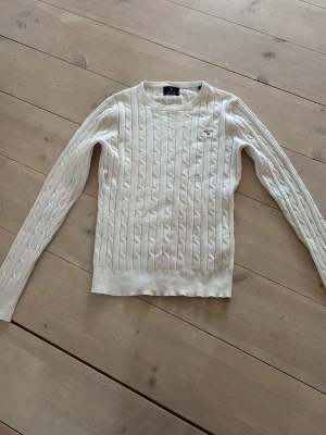 Vit kabelstickad tröja från Gant - Kabelstickad vit tröja från Gant med rund halsringning och broderad logga på bröstet. Tröjan har långa ärmar och ribbade muddar vid ärmslut och nederkant. Perfekt för dig som gillar klassisk och stilren design.