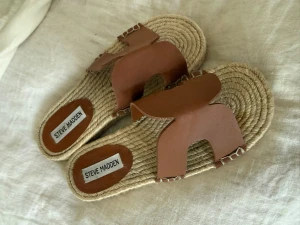 Bruna sandaler från Steve Madden - Snygga sandaler från Steve Madden med flätad sula i naturmaterial och breda bruna remmar i läder. Öppen tå och slip-in modell med dekorativa sömmar längs kanterna. Perfekta för en avslappnad och trendig look.