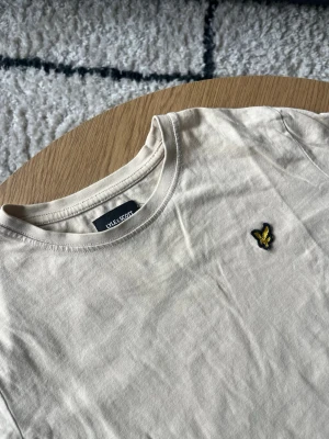 Beige Lyle & Scott T-shirt - Hejsan✌️ Säljer en nygg beige t-shirt från Lyle & Scott med klassisk rund halsringning och korta ärmar🤩 På bröstet sitter den ikoniska broderade loggan i gult och svart🪡 Tillverkad i mjuk bomull för en skön känsla och enkel stil👕