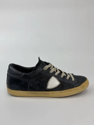 Philippe Model svarta sneakers - Svarta sneakers från Philippe Model med beige sula och vita skosnören. Skorna har en ikonisk vit sköld på sidan och är tillverkade i läder med mockadetaljer. Klassisk låg modell som passar till många olika stilar.