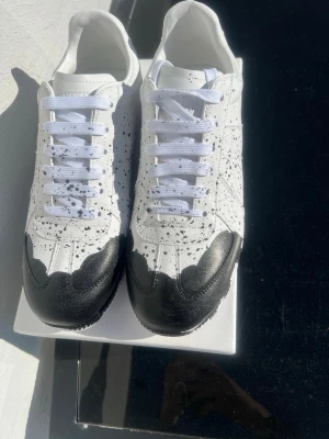 Vita Maison margielas med svart färg - Unika vita sneakers med svart splash-mönster och svart tåparti. Skorna har vita skosnören och en låg silhuett. Materialet ser ut att vara läder eller konstläder. Perfekta för dig som vill sticka ut med en kreativ och modern look.
