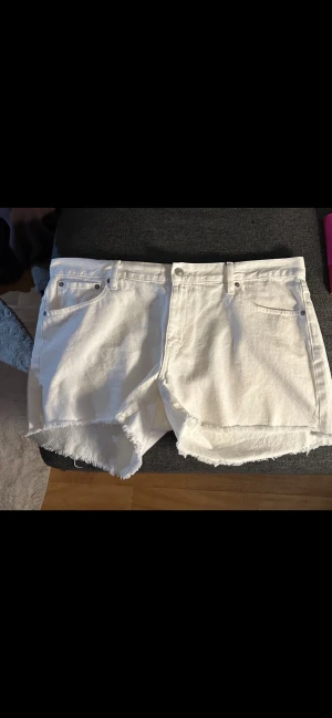 Vita jeansshorts från 157 med rå kant - Säljer ett par vita jeansshorts från 157 med råa, fransiga kanter. Klassisk femficksmodell med knapp och dragkedja framtill. Perfekta för varma dagar och enkel att matcha med allt. Snygg och avslappnad stil.
