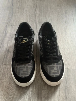 Svarta Fendi sneakers med FF-mönster - Snygga svarta sneakers från Fendi med klassiskt FF-mönster i grått och svart. Skorna har vita sulor, svarta skosnören och detaljer i mocka och textil. Perfekta för dig som gillar exklusiv streetstyle och vill sticka ut med ett lyxigt märke.