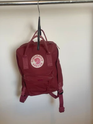 Vinröd Fjällräven Kånken mini  ryggsäck - Vinröd klassisk Fjällräven Kånken ryggsäck med ikonisk rund logotyp framtill. Ryggsäcken har justerbara axelremmar, dubbla handtag upptill och en dragkedja som öppnar huvudfacket. Tillverkad i slitstarkt syntetmaterial.