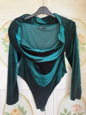 Grön sammetsbody från NLY One - Snygg grön sammetsbody från NLY One med lång ärm och draperad halsringning. Perfekt för dig som vill sticka ut med en lyxig känsla. Materialet är mjukt och glansigt.
