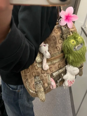 Beige handväska med guldiga detaljer - Cool beige handväska från Guess med guldiga spännen och detaljer. Väskan har ett monogrammönster och är dekorerad med flera söta och roliga nyckelringar, bland annat mjukisdjur och en rosa blomma. Perfekt för dig som vill sticka ut med din stil. Y2k mcbling streetwear
