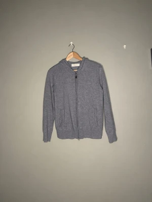 Kashmir tröja från Sayless - Kashmir tröja från Sayless i storlek XS. En stilren mörkgrå hoodie med dragkedja framtill och två fickor. Hoodien har en klassisk huva och är tillverkad i ett mjukt, stickat material som känns skönt mot huden. Perfekt för dig som gillar en enkel och clean look.