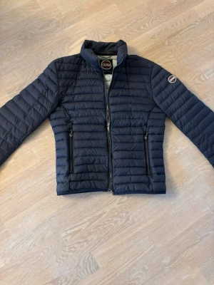 Mörkblå pufferjacka från Colmar - Snygg mörkblå pufferjacka från Colmar med quiltad design och två dragkedjeförsedda fickor framtill. Jackan har hög krage och en diskret logotyp på ärmen. Insidan är ljusgrå och jackan stängs med dragkedja. Perfekt för kalla dagar.