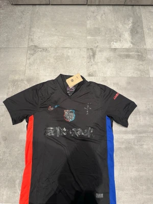 Svart FC Barcelona matchtröja Nike x travis - Snygg svart FC Barcelona matchtröja från Nike med klubbmärke och Nike-logga på bröstet. Tröjan har röda och blå ränder på sidorna, korta ärmar och tryck på både fram- och baksida. Tillverkad i lätt och ventilerande funktionsmaterial.