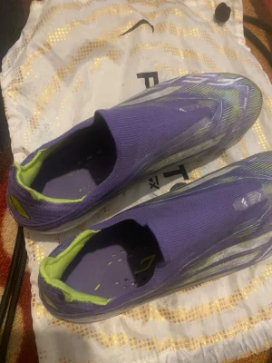 Lila Adidas X Speedflow fotbollsskor - Säljer ett par lila Adidas X Speedflow fotbollsskor med neongröna detaljer och vit sula. Skorna har en strumpliknande passform utan snörning och är designade för maximal fart på planen. Perfekta för dig som vill sticka ut med både stil och prestanda.