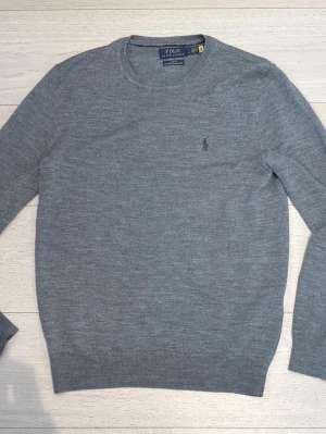 Grå merinoulltröja Polo Ralph Lauren - Snygg grå stickad tröja från Polo Ralph Lauren i slim fit-modell. Tillverkad i tvättbar merinoull och har rund hals samt diskret broderad logga på bröstet. Perfekt för en stilren och bekväm look.