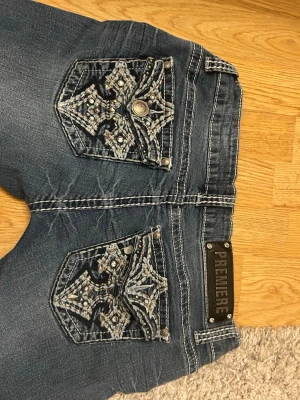 Blå jeans med broderade bakfickor - Snygga blå jeans från Premiere med unika broderade detaljer och nitar på bakfickorna. Jeansen är omsydda till ytterligare lägre midja. De passar bra på mig som är 170. Säljer då de är lite stora i midjan 
