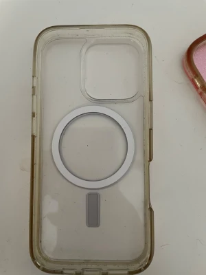 Transparent MagSafe mobilskal - Säljer ett genomskinligt mobilskal med MagSafe-kompatibilitet. Skalets kanter är lätt gulnade och det har en tydlig cirkel för MagSafe på baksidan. Passar iPhone-modeller med större kameralins. Tillverkat i tålig plast.
