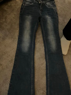 Mörkblå bootcut jeans med broderi - Säljer ett par mörkblå bootcut jeans med snygga slitningar framtill och broderade bakfickor med vita detaljer. Klassisk femficksmodell och knappgylf. Jeansen har kontrastsömmar och är tillverkade i denim med lite stretch för skön passform.