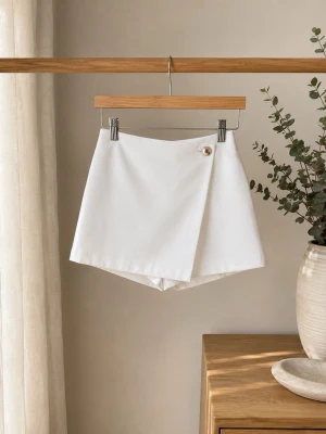Vita skorts med omlott och knapp - Stilrena vita skorts med omlott-design framtill och dekorativ knapp i midjan. Skortsen har en rak passform och en diskret ficka bak. Perfekt för en fräsch och trendig look. Materialet ser ut att vara mjukt och lätt, troligen bomullsblandning.