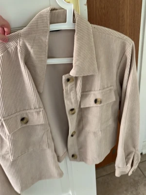 Beige overshirt i manchester - Snygg beige overshirt i manchestertyg med stora bröstfickor och knappar framtill. Overshirten har klassisk krage och långa ärmar med knapp vid ärmslut. Perfekt för lager-på-lager och ger en chill vibe till din outfit.