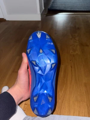 Adidas Predator blå/röda fotbollsskor - Säljer ett par Adidas Predator fotbollsskor i blått och rött med coola mönster och klassiska tre ränder på sidan. Skorna har en slitstark sula med fasta dobbar och är tillverkade i syntetmaterial för bra grepp och kontroll på planen.