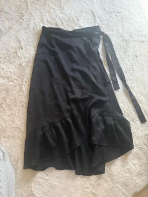 Svart omlottkjol med volang - Svart omlottkjol med volangkant och knytband i midjan. Kjolen har en elegant och flowy design som ger en snygg siluett. Perfekt för dig som gillar stilrena plagg med en twist.