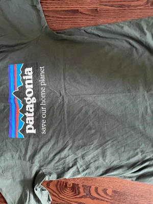Grön Patagonia t-shirt med tryck - Snygg grön t-shirt från Patagonia med stort färgglatt tryck och texten 'save our home planet' på ryggen. Framtill finns en liten Patagonia-logga. Klassisk passform och rund hals. Perfekt för en avslappnad stil.