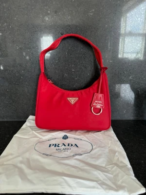 Röd axelremsväska  - Snygg röd handväska. Ej äkta i klassisk baguette-modell. Väskan är tillverkad i nylon med silvriga metalldetaljer och har trekantslogga framtill. Axelremmen är bred och bekväm, och väskan kommer med dustbag