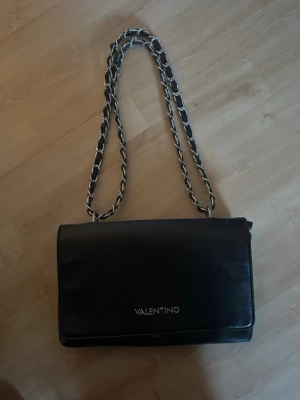 Svart axelväska från Valentino - Snygg svart axelväska från Valentino med silverfärgad kedjerem och logga framtill. Väskan har en stilren, rektangulär form och är tillverkad i slätt material som ger en lyxig känsla. Perfekt för dig som gillar klassisk och trendig design. Har ett skav på märket bild 3 och även lite sliten på bandet men inget som syns vid användning💗