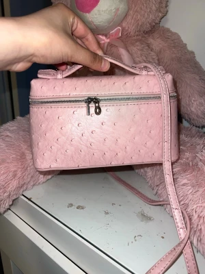 Ljusrosa handväska med struktur - Söt ljusrosa handväska i boxig form med struktur som påminner om struts. Väskan har ett handtag upptill, lång axelrem och silverfärgad dragkedja. Perfekt för dig som gillar gulliga accessoarer och vill sticka ut lite extra.