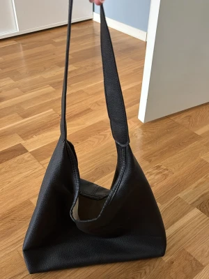Svart axelväska i skinn - Stilren svart axelväska i mjuk skinnimitation med minimalistisk design och bred axelrem. Väskan har en slouchy form och passar perfekt för dig som gillar en enkel men trendig look. Perfekt storlek för att få plats med allt du behöver till vardags.