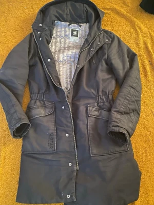Svart parkas från G-Star RAW - Svart parkas med huva från G-Star RAW. Jackan har stora fickor framtill, dragkedja och knappar, samt quiltat foder i grått. Snygg, rak modell med markerad midja och diskret logga på ena fickan. Perfekt för kyliga dagar.