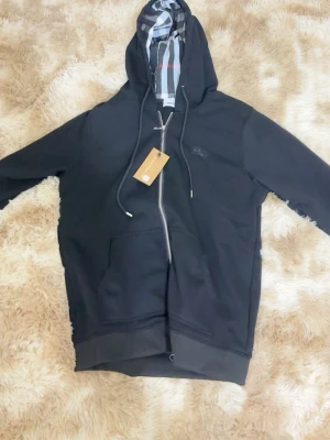 Svart hoodie från Burberry med dragkedja - Svart hoodie från Burberry med hel dragkedja, klassisk känguruficka och justerbar huva. Insidan av huvan har Burberrys ikoniska rutmönster i svart, vitt och rött. Broderad logga på bröstet. Perfekt för en stilren och avslappnad look.