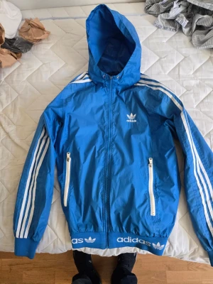 Blå Adidas vindjacka med huva - Fin adidas jacka, litet hål under armen som lätt sys igen. 