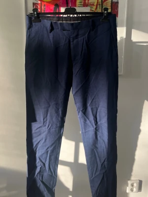 Mörkblå kostymbyxor i rak modell - Snygga mörkblå kostymbyxor med rak passform och klassisk design. Byxorna har hällor för bälte, två bakfickor med knapp och är tillverkade i ett lätt tyg som ger en dressad look. Perfekta för en stilren outfit. Tillhörande kavaj i nästkommande annons. 