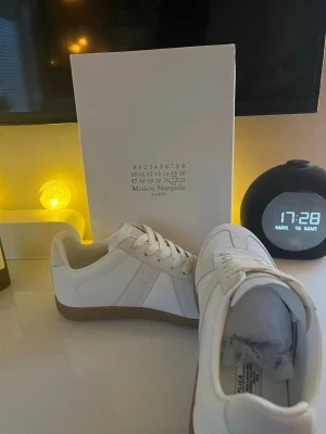 Maison Margiela vita sneakers kvitto finns  - Snygga vita sneakers från Maison Margiela med klassisk design. Skorna har ovandel i vitt läder och mocka, samt en brun gummisula. Stilrena detaljer och vita skosnören gör dem perfekta för en clean look. Märkeslogga på plösen och insidan.