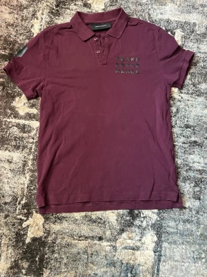 Vinröd pikétröja Peak Performance - Snygg vinröd pikétröja från Peak Performance med klassisk krage och knappar framtill. Diskret logga på bröstet och korta ärmar. Perfekt till jeans eller shorts för en clean look.