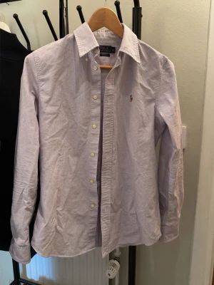 Ljuslila skjorta från Polo Ralph Lauren - Klassisk lite blå och lila skjorta från Polo Ralph Lauren med broderad logga på bröstet. Skjortan har lång ärm, knappar framtill och en tidlös krage. Perfekt för dig som gillar stilrena och snygga plagg.