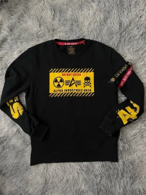 Svart sweatshirt Alpha Industries - Cool svart sweatshirt från Alpha Industries med stora gula tryck och varningsskylt på bröstet. Tröjan har rund hals, ribbade muddar och detaljer som röd tag och dragkedja på ärmen. Perfekt för dig som gillar streetwear och tydliga statement-plagg.