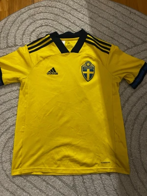 Svenska landslagströja Adidas gul - Gul fotbollströja från Adidas med Sveriges landslagsemblem på bröstet och svarta detaljer. Tröjan har korta ärmar, svart krage och tre svarta ränder på axlarna. Tillverkad i lätt och ventilerande material med AEROREADY-teknologi.