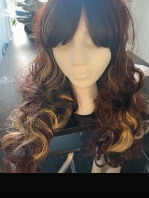Lockig peruk med bruna och blonda slingor - Snygg lockig peruk i syntetmaterial med djupbrun bas och breda blonda slingor. Långt hår med mycket volym och naturligt fall. Perfekt för att förändra din look snabbt och enkelt.