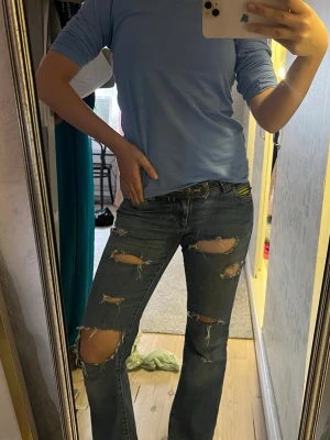 Low waist, blå slitna bootcut jeans - Säljer ett par blå bootcut jeans med flera stora slitningar och hål på benen. Jeansen har klassiska bakfickor med dekorativa sömmar och gula detaljer vid hällorna. Perfekta för en avslappnad och trendig look. Sitter välldigt low waist. W/27 L31 ( skulle jag tro) skickar mått i dm☺️