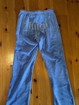 Blå velourbyxor från Juicy Couture - Blå mjukisbyxor i velour från Juicy Couture med strassdetaljer och texten 'Juicy' på baksidan. Byxorna har resår i midjan, raka ben och dragkedjeförsedda fickor framtill. Perfekta för en avslappnad och trendig look.