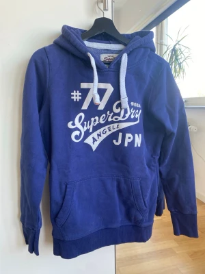 Blå Superdry hoodie med tryck - Snygg blå hoodie från Superdry med vitt tryck framtill och stora fickor. Klassisk huva med dragsko och ribbade muddar. Perfekt för en avslappnad streetstyle-look.