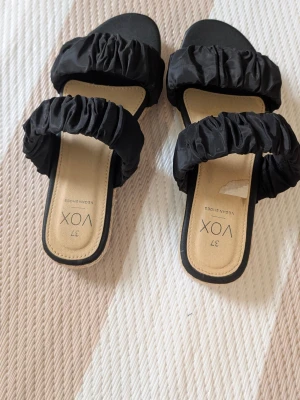 Svarta sandaler från VOX med resårband - Säljer ett par svarta sandaler från VOX med två breda, rynkade resårband över foten. Sandalerna har fyrkantig tå och beige innersula med svart kant. Sulan är låg och har en flätad detalj på sidan. Perfekta för en trendig och avslappnad look.