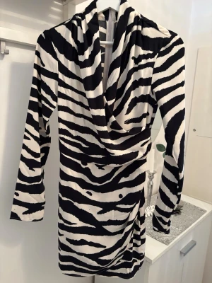 Zebramönstrad kort klänning - Snygg kort klänning med zebramönster i svart och vitt. Klänningen har lång ärm, omlottkrage och markerad midja. Perfekt för dig som vill sticka ut med en trendig look.