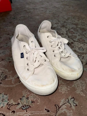 Vita tygsneakers från Novesta - Klassiska vita sneakers från Novesta i canvas med lågt skaft och vita snören. Skorna har en diskret logotyp på sidan och en robust vit gummisula. Perfekta för en clean och avslappnad stil.